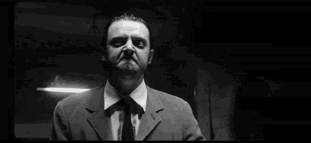 a man in a suit and tie is standing in a dark room with his hands folded in front of him .