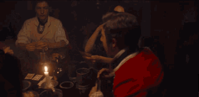 a man in a red coat sits at a table with candles and playing cards