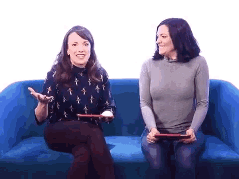 two women are sitting on a blue couch holding tablets and smiling .