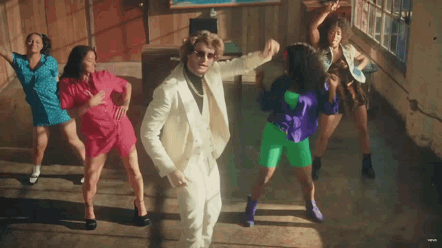 a man in a white suit is surrounded by a group of women dancing