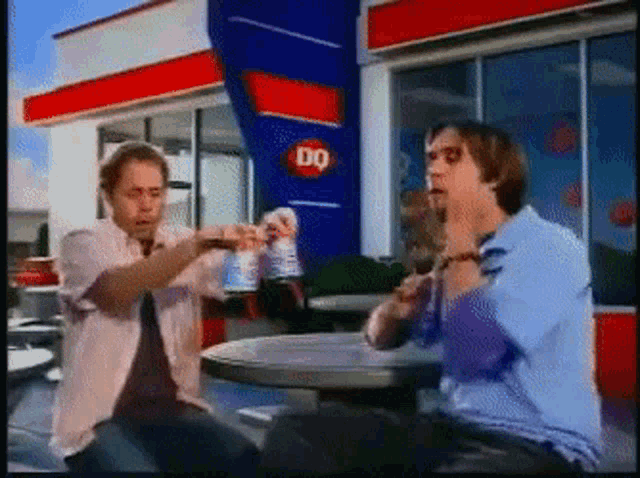 two men are sitting at a table in front of a building that says dq
