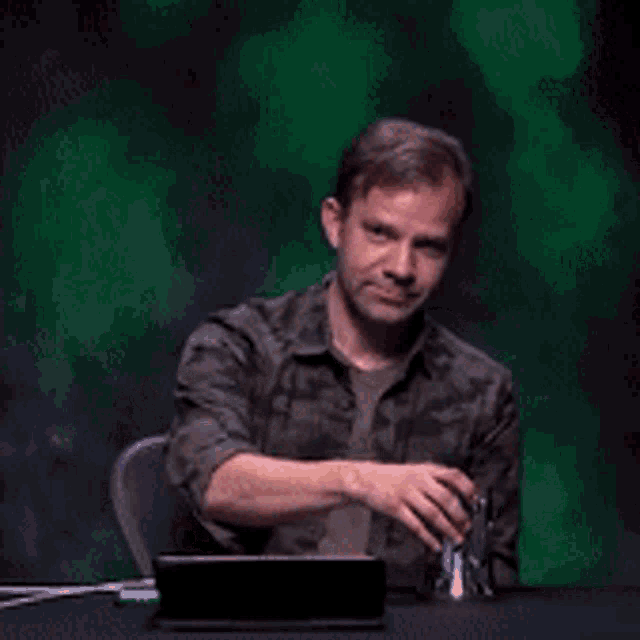 a man in a camo shirt is sitting at a table laughing while holding a cup .