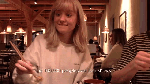 a woman in a white sweatshirt is holding chopsticks in front of a sign that says is 150,000 people over four shows