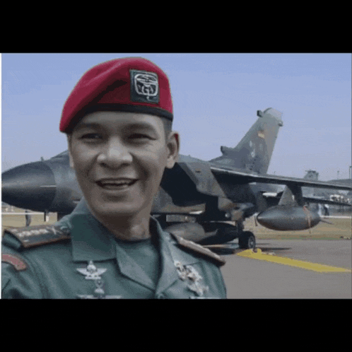 a man wearing a red beret with the letter y on it smiles in front of a fighter jet