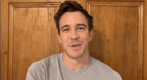 a man in a blue t-shirt is smiling in front of a wooden wall .