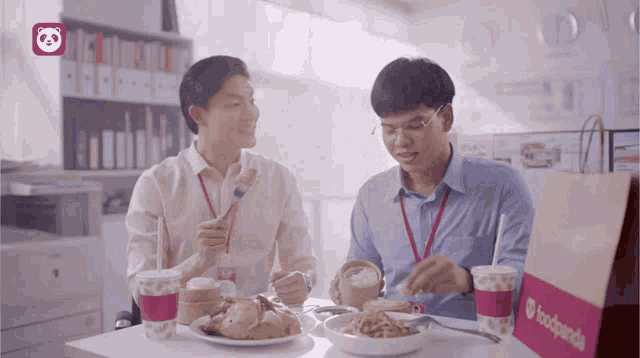 two men are sitting at a table eating food and drinking from a bag that says foodpanda