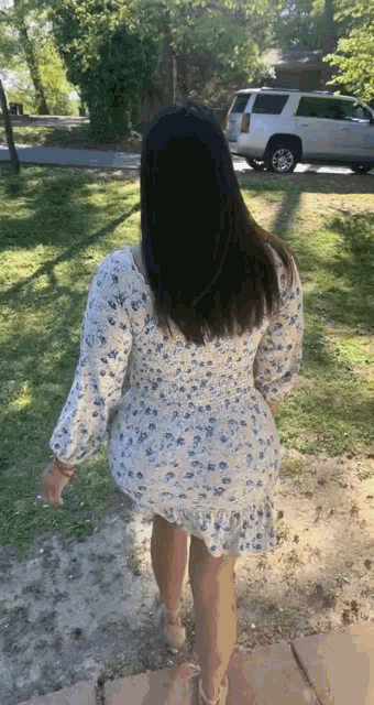 a woman in a white floral dress is walking down a sidewalk in front of a white suv .