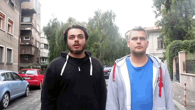 two men standing next to each other with one wearing a blue shirt and the other a gray hoodie