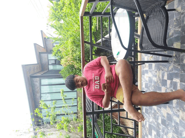 a man sits on a balcony wearing a red shirt that says " dad "