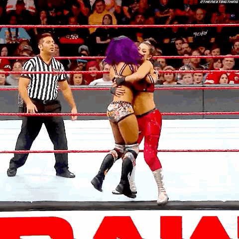 a woman with purple hair is hugging another woman in a wrestling ring while a referee looks on