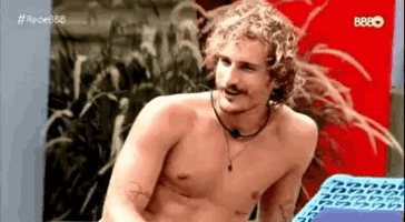 a shirtless man with curly hair and a mustache is sitting on a purple block .