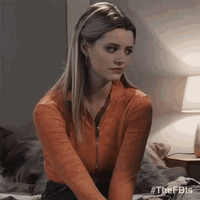 a woman is sitting on a bed wearing an orange sweater and looking at the camera .