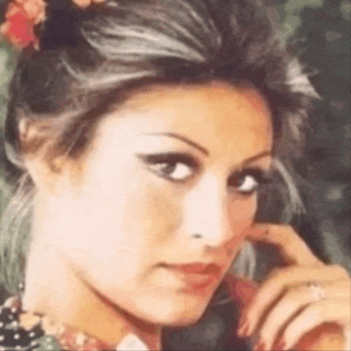 a woman with a flower in her hair is looking at the camera with her hand on her face .