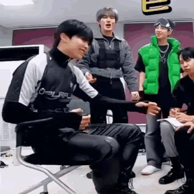 a group of young men are sitting around a table in a room . one of the men is wearing a green vest .