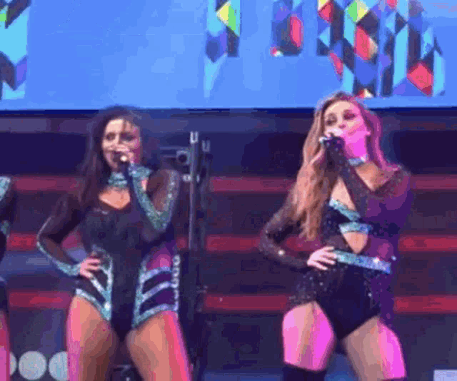 two women singing into microphones on a stage with a large screen behind them