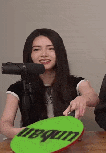 a woman is sitting in front of a microphone holding a green sign that says ' uegum ' on it .