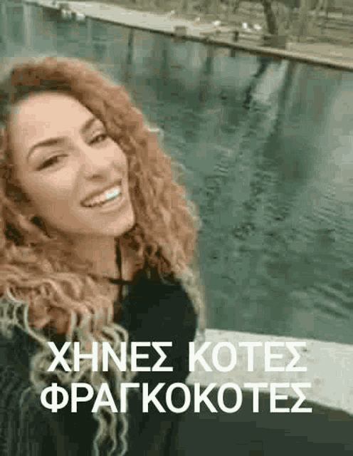 a woman with curly hair is smiling in front of a body of water in greek .