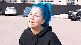 a woman with blue hair is wearing a black hoodie and smiling in a parking lot .
