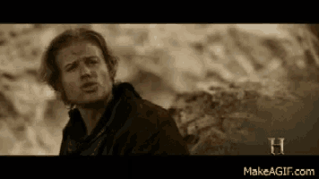 a man in a brown jacket is standing in the sand and talking to someone .