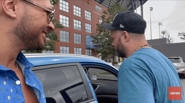 two men are talking in front of a blue car with a subscribe button on the bottom
