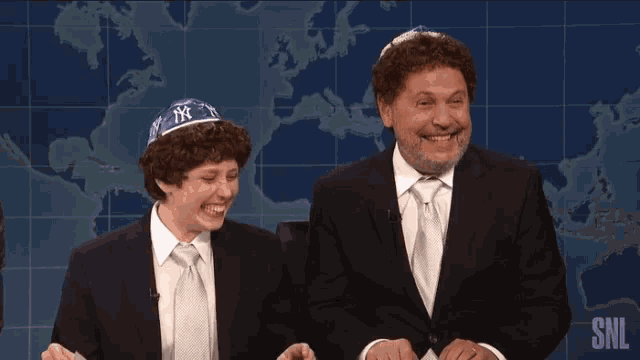 a man in a suit and tie laughs next to another man in a yarmulke on a snl show
