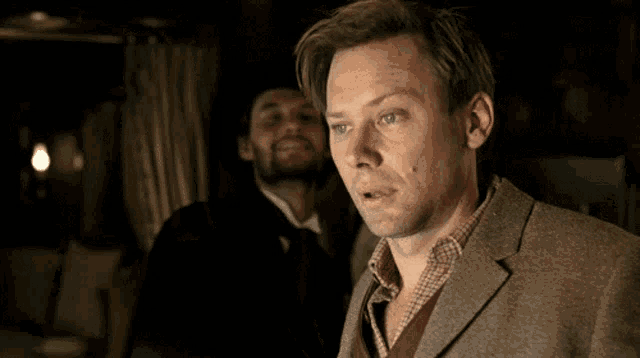 a man in a suit stands in front of another man in a hat