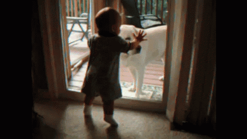 a little girl is standing in front of a door reaching out towards a dog