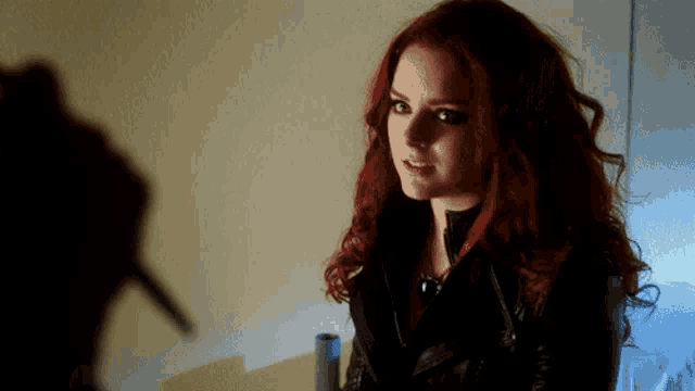 a woman with red hair and a black jacket looks at the camera