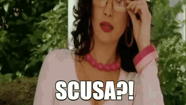 a woman wearing glasses and a pink necklace is talking on a cell phone and says scusa ?