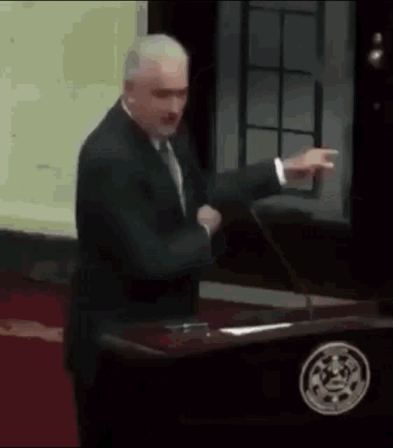 a man in a suit and tie stands at a podium pointing at something