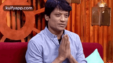 a man in a blue shirt is sitting on a red couch with his hands folded in prayer .