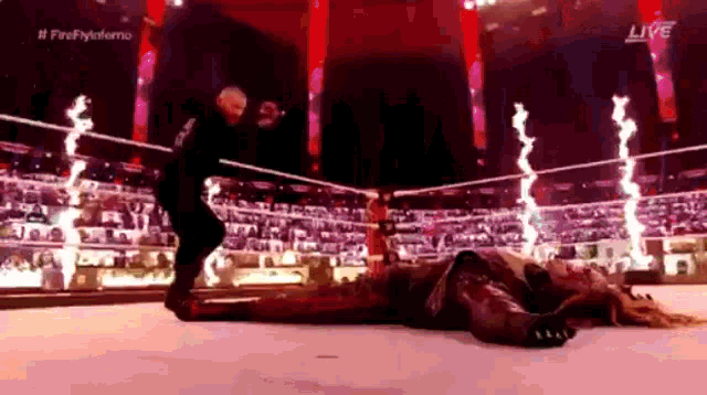 a man is standing next to a man laying on the ground in a wrestling ring with fireworks behind him .
