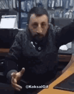a man with a beard is sitting at a desk with his arms outstretched and making a funny face .