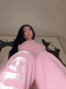 a woman wearing pink sweatpants and a pink hoodie is sitting on a bed .