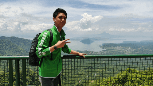 a young man wearing a green adidas jacket stands on a fence overlooking a lake