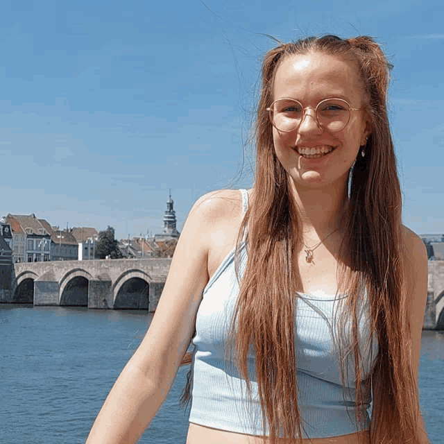 a woman wearing glasses and a blue tank top smiles in front of a bridge over a body of water