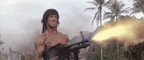 a shirtless man is holding a machine gun in a field with palm trees in the background .