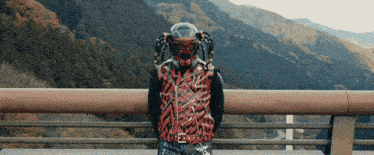 a man wearing a helmet and a red jacket stands on a bridge overlooking mountains