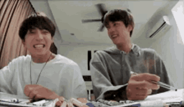 two young men are sitting at a table laughing while holding pens .
