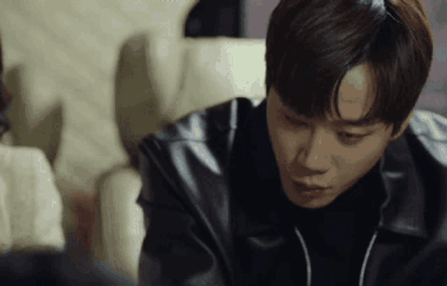 a man in a black leather jacket is sitting on a couch looking down .