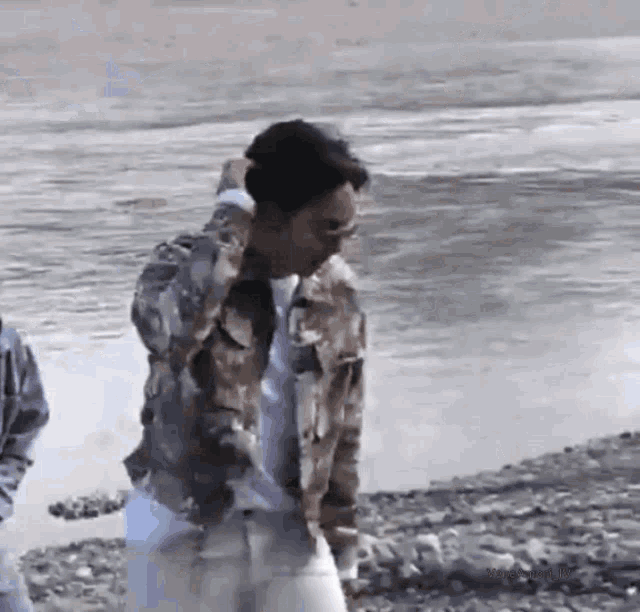 a man in a camouflage jacket is standing on a rocky beach next to a body of water .