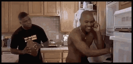 a shirtless man is talking on a cell phone in a kitchen while another shirtless man holds a football .