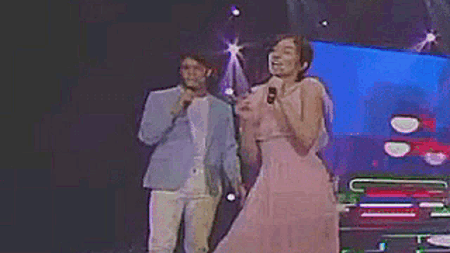 a woman in a pink dress is singing into a microphone while standing next to a man in a blue jacket .