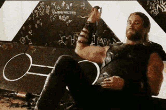a man with a beard is sitting on the floor in front of a chalkboard with the letter o on it