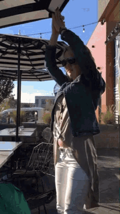 a woman wearing sunglasses and a denim jacket holds her hands in the air