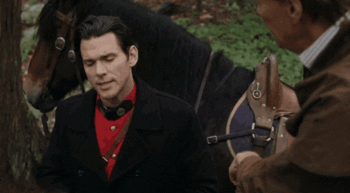 a man in a black coat and red shirt stands next to a horse