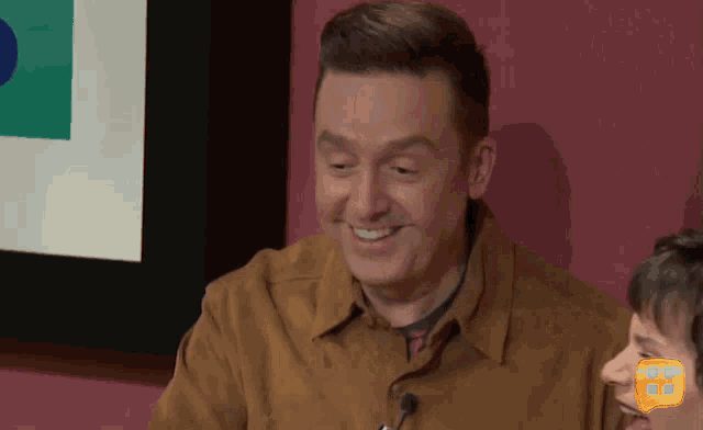a man in a brown shirt smiles while sitting in front of a microphone