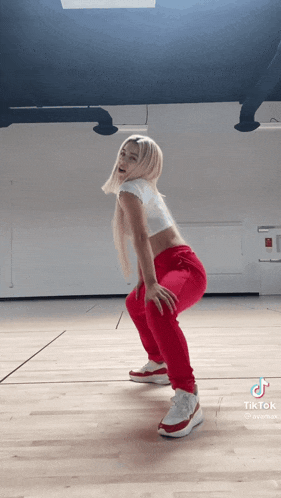 a woman wearing red sweatpants and white sneakers is squatting on a wooden floor