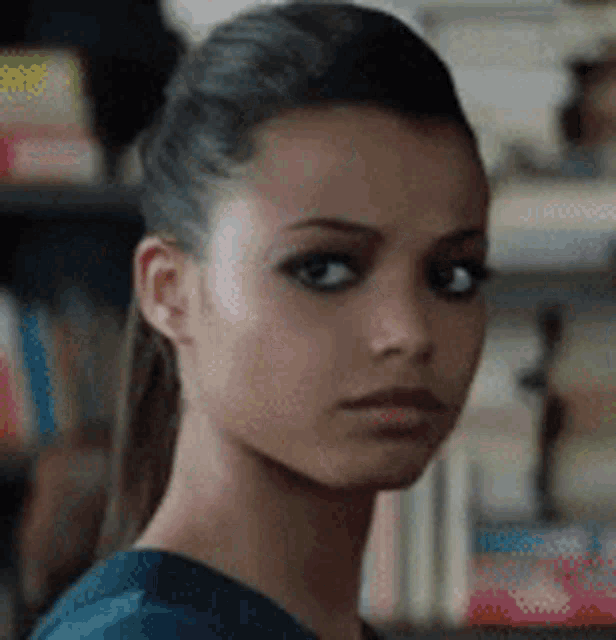 a close up of a woman 's face with a ponytail and a bookshelf in the background .