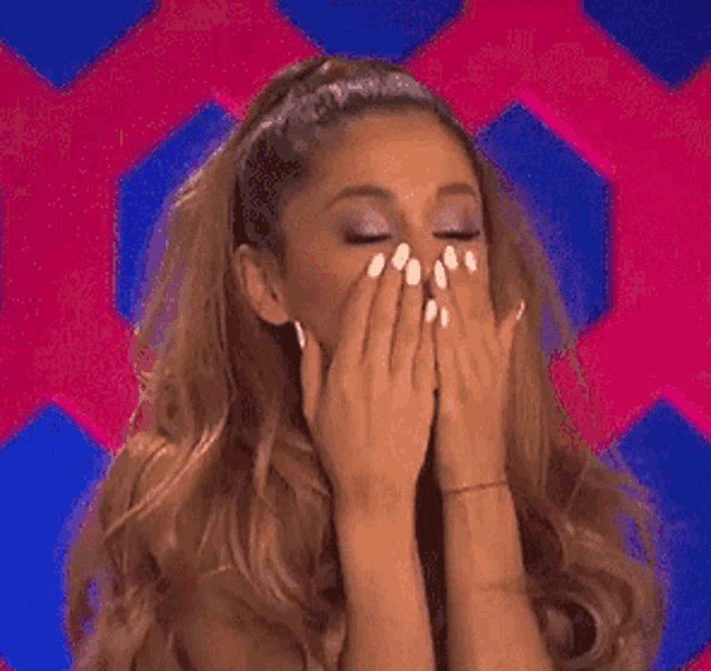 ariana grande is covering her mouth with her hands while standing in front of a pink and blue background .